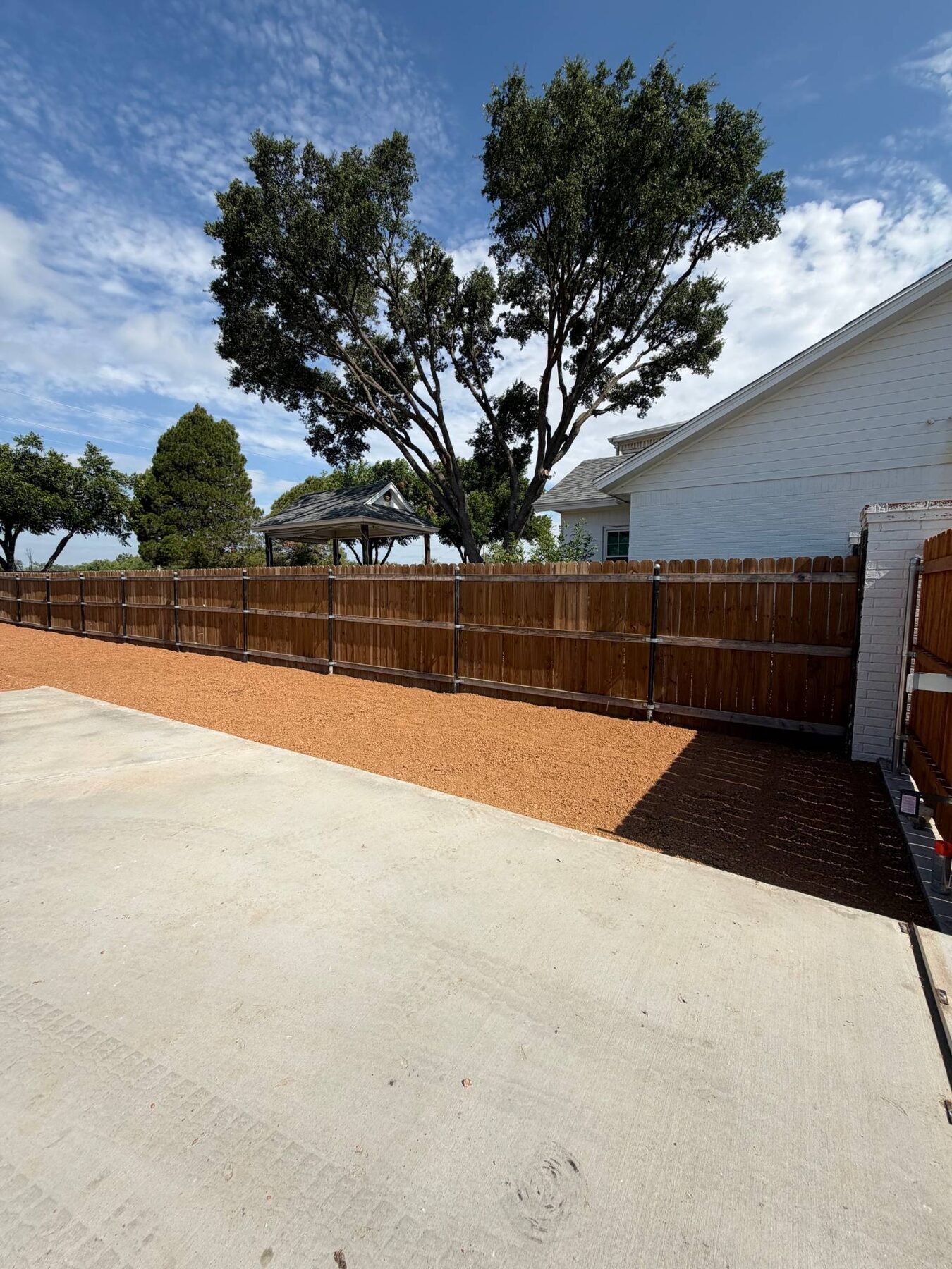 Red rock ground cover along a wood privacy fence beside a concrete driveway, with a large oak tree behind